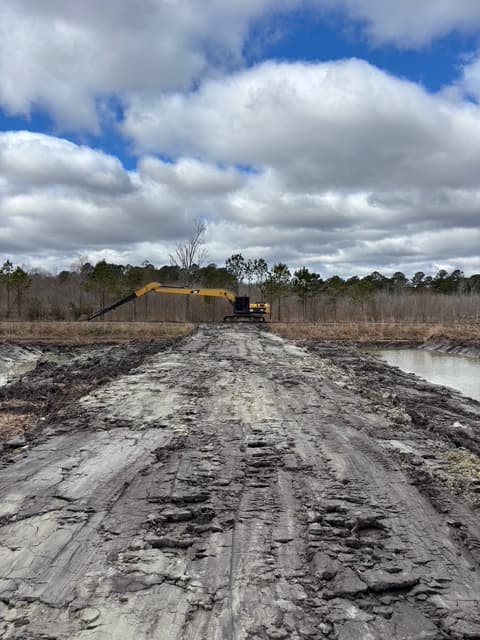 Yellow long-reach excavator at the end of a muddy, rutted path under a cloudy sky.