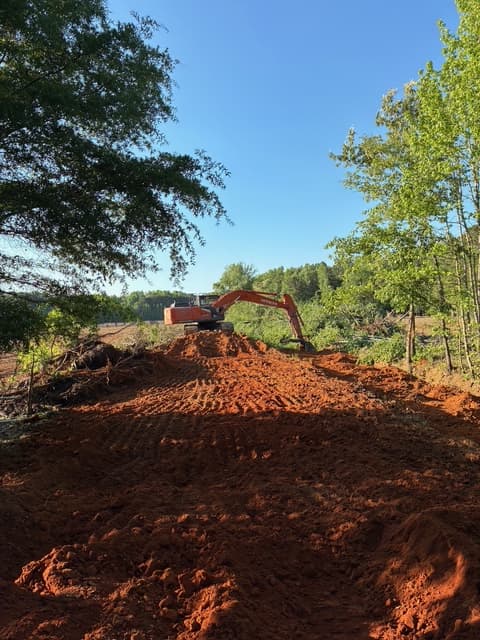 Orange excavator sits on a large mound of red clay surrounded by green trees.